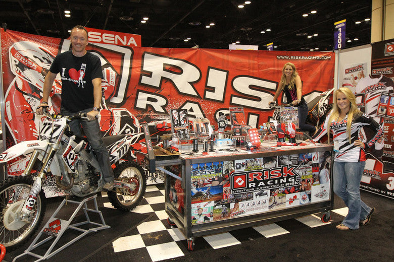 Risk Racing booth at the SENA show full of products including RR owner James Burry posing on a dirt bike sitting on a RR1 stand