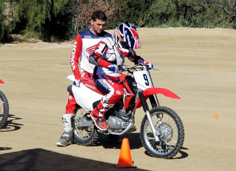 Young dirt bike rider learning the basics of riding a dirt bike under the guidance of an older and more experienced motocross rider. The young rider is sitting on the bike in a ready position as the trainer holds the bike up and balanced.