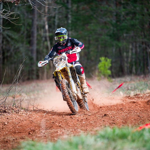 Motocross rider wearing risk racing J.A.C. V3 Roll-off goggles while accelerating up hill. The rider and bike are very dirty from the race and the rider has used the roll-off system to maintain clear vision.