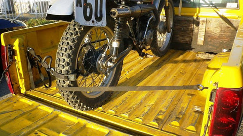 Moto vintage attachée dans un lit de camion jaune avec quatre sangles à cliquet marron. À attaché au guidon, et deux autres attachés à la roue arrière l'empêchant de rouler vers l'arrière.
