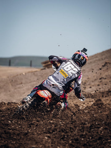 Motocross rider on a 2023 crf450r with red plastics wearing white black and pink gear leaning to the right through a loose corner with dirt flying off of the rear tire.