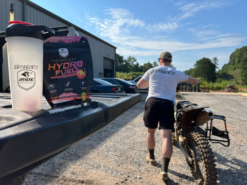 Piloto de motocross empujando su bicicleta con su RISK Racing A.T.S. soporte apoyado sobre su pedal de arranque para su transporte. El conductor tiene una bolsa de iRide Supplements Hydro Fuel y una botella agitadora RISK Racing/iRide en el portón trasero de su camión para obtener electrolitos en su sistema antes de viajar.