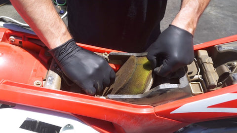 Changing/cleaning the air filter on this Honda dirt bike. the person working on this bike had to remove the seat to access the air filter. They have removed the wing screw that attaches/seals the filter and filter cage to the dirt biek and is removing the filter form the bike.