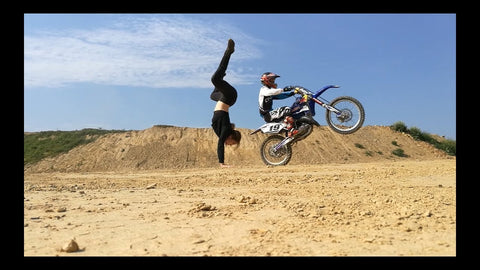 One person wheelieing a dirt bike while another person performs a yoga handstand move at a dirt bike track. 