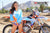 Plano medio de la modelo de moto Alliyah de febrero con una camiseta de MX Risk Racing azul y naranja y pantalones de bikini azules de pie frente a una moto de cross KTM n.º 277 en una pista de MX con montañas al fondo.