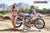 Wide shot of February Moto Model Alliyah in a bikini standing in front of a KTM dirt bike #277 sitting on a Risk Racing ATS MX stand at a MX track with mountains in background. Her left arm across torso, right hand touching her hair.