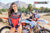 Close up of February Moto Model Alliyah tugging her Black & Red Risk Racing MX Jersey down almost covering her blue bikini bottoms standing in front of a KTM dirt bike on an ATS stand at a MX track with mountains in background.