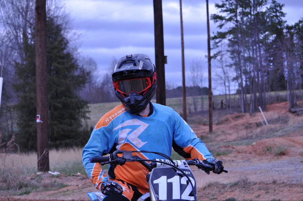 mx num 112 person sitting on dirt bike wearing blue and orange risk racing jersey land n tress in BG