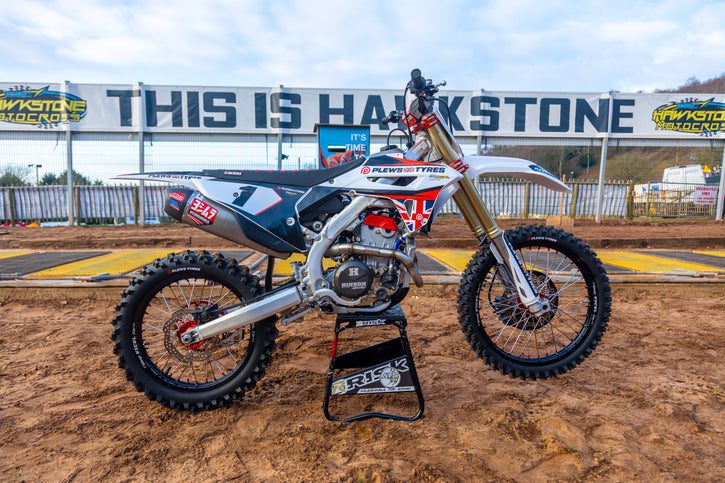 motocross bike wearing Plews tires sitting on a Risk Racing A.T.S. stand at an MX track with a giant sign in the background that reads: this is hawkstone