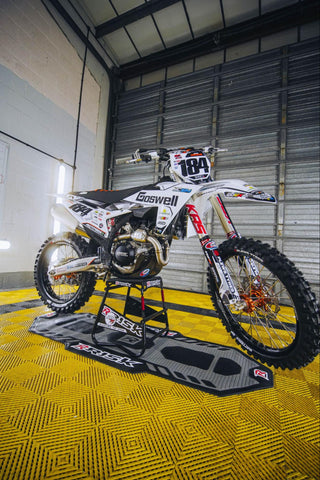 Brand new 2024 KTM 450 motocross race bike with white plastics and white and black graphics resting on top of a RISK Racing A.T.S. Stand to get both tires off the ground inside of a climate controlled garage with a yellow floor. Also pictured is a RISK racing floor mat to keep the garage tidy and make the set up look professional. The floor mat is underneath the stand and is colored black and grey, with red RISK Racing logos.
