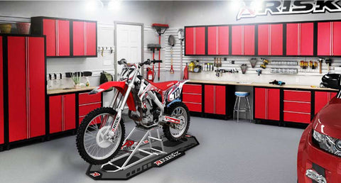 dirtbike on a risk racing pitmate and ride-on lift inside of a well lit garage courtesy of a trilight garage light. The shop has many well organized work benches and tool/part storage areas.