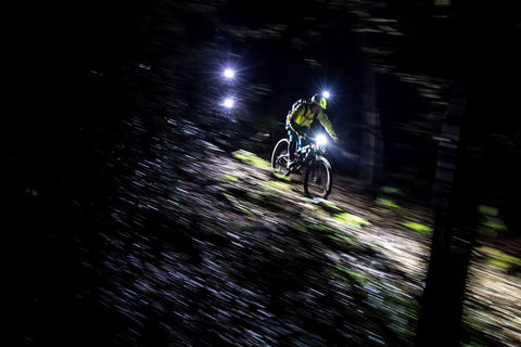 Two riders descending together on a single track mountain bike trail. each rider has two lights. One light is a headlamp, another light is mounted on the handlebars.
