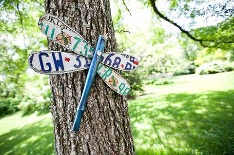 A metal yard art dragonfly made of old license plates is affixed to a tree trunk.