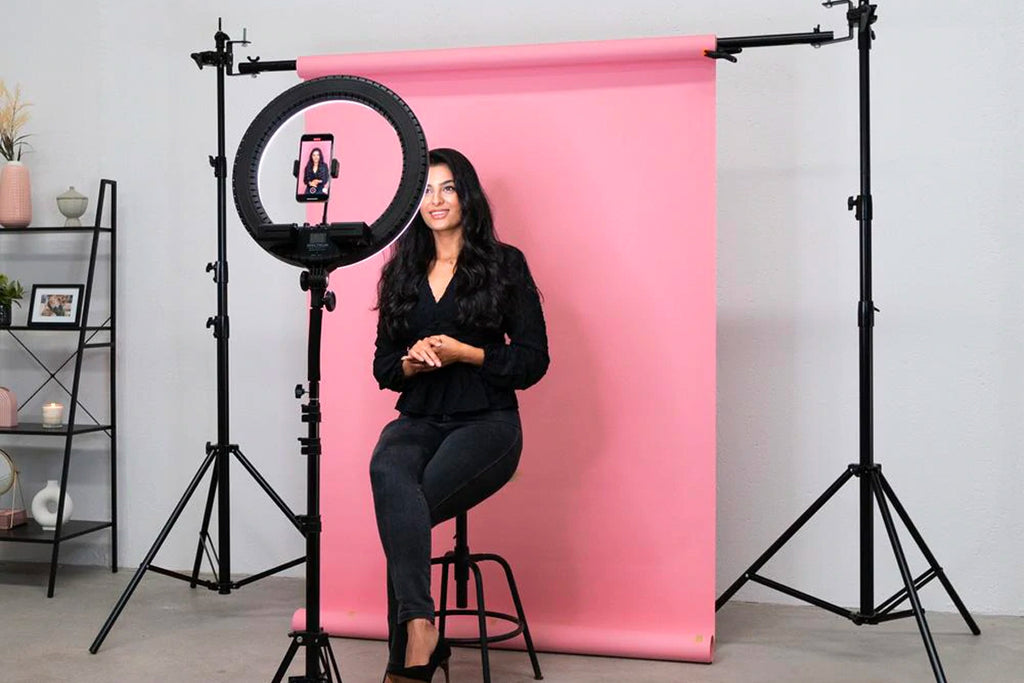 Woman sitting on stool in from of pink paper backdrop using phone mounted on a ring light