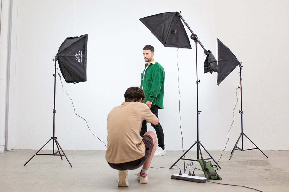 an image of a photographer a man kneeling down to take a full length shot of a male fashion model in a white backdrop with a three-point lighting setup.
