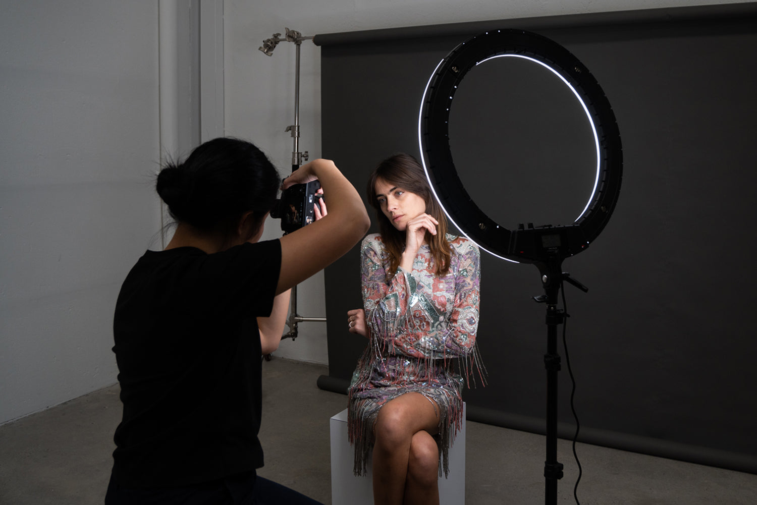 a female photographer taking a photo of a fashion model sitting on a stool with a one-point lighting setup using ring light.