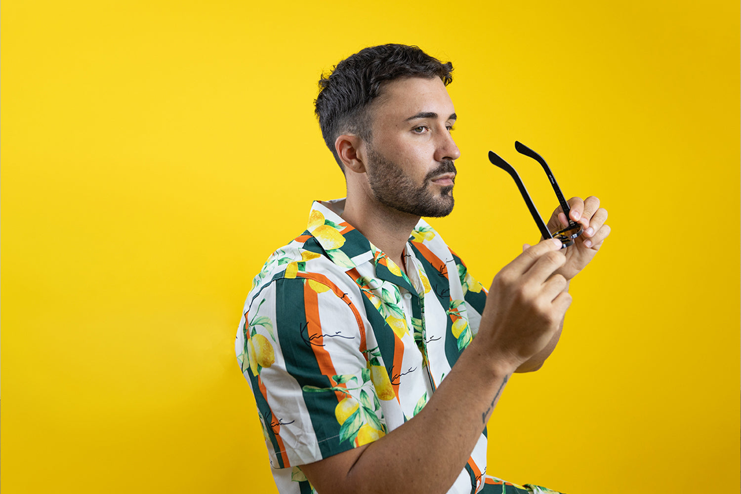 a medium length shot of a fashion model wearing a tropical polo and holding a pair of sunglasses in front of his face.