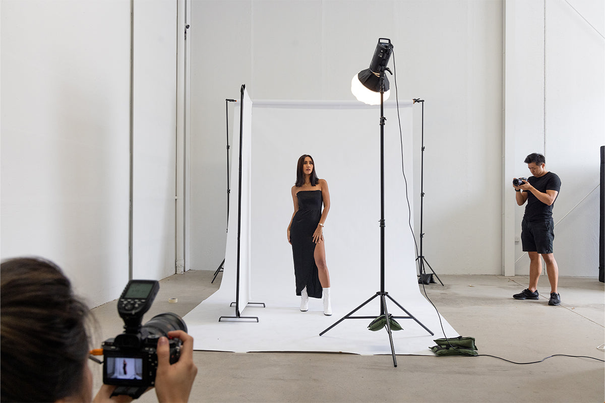 A wide shot photograph of a professional photoshoot, showing a Godox DP600III lantern softbox in the frame. The softbox is positioned to the right of the model, casting a warm, diffused light on her face and surroundings. The model is standing in front of a white backdrop, while in the background, two photographers are visible, one holding a camera and taking the model's picture, and the other observing. The lantern design of the softbox creates a unique circular catchlight in the model's eyes. The softbox, photographers, and model all give a glimpse into a professional photoshoot environment.
