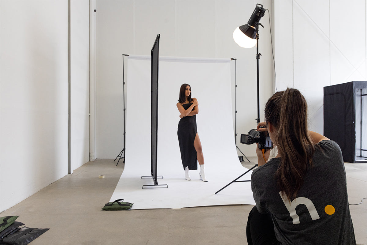 A wide shot photograph of a professional photoshoot, showing a Godox DP600III lantern softbox in the frame. The softbox is positioned to the right of the model, casting a warm, diffused light on her face and surroundings. The model is standing in front of a white backdrop, while in the foreground, a photographer is seen holding a camera and taking the model's picture
