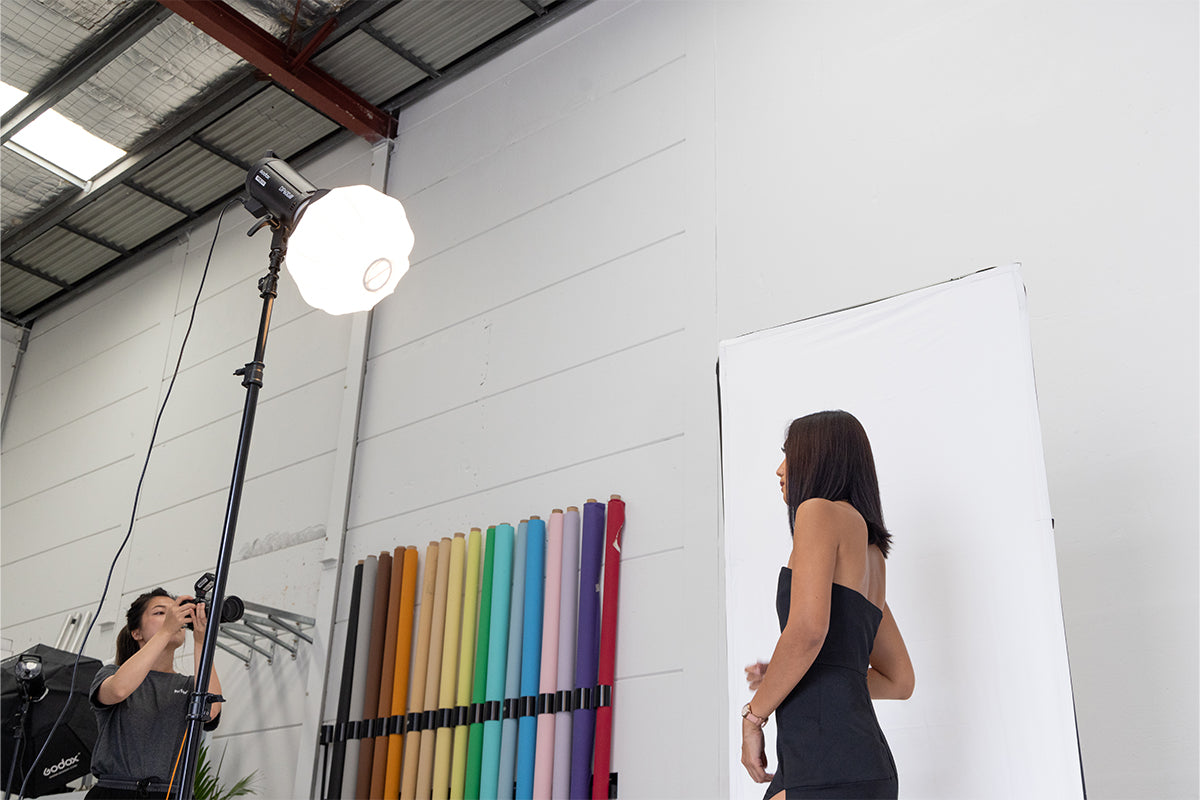  A photograph of a model being illuminated by a Godox DP600III lantern softbox during a photoshoot. The softbox is positioned right above the frame casting a soft, diffused light on the model's face and surroundings. In the foreground, a photographer is seen holding a camera and taking the model's picture. The lantern design of the softbox creates a unique circular catchlight in the model's eyes, adding depth and dimension to the image.