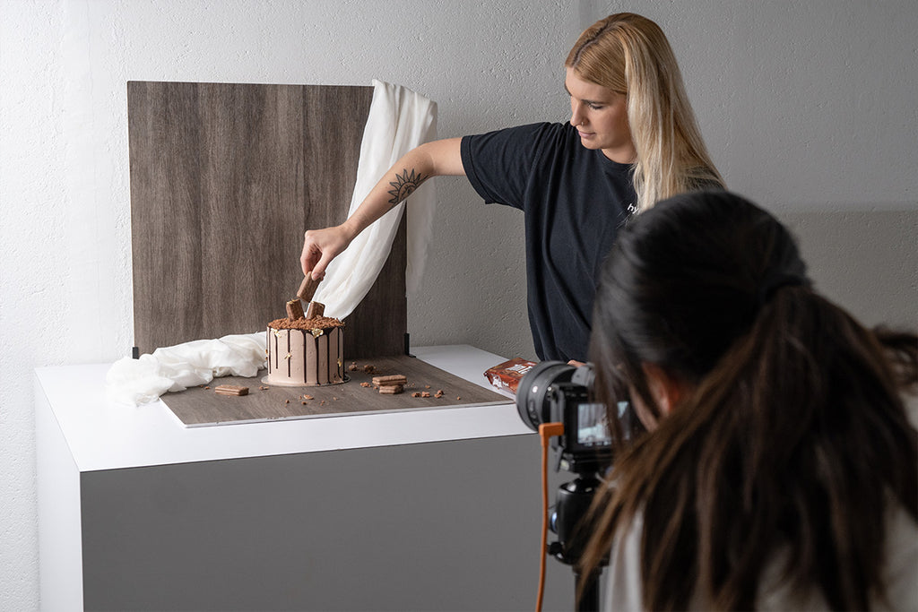 Photographer taking a photo of a chocolate cake while female assistant holds a Tim Tam above the cake