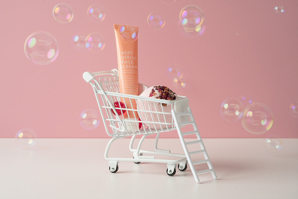 Skincare product and bath bomb positioned in a mini shopping trolley which is in front of a pink backdrop 