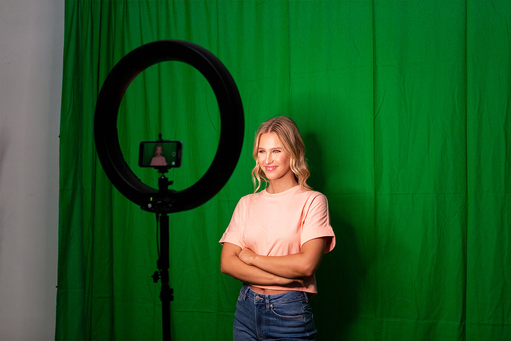 Smiling woman standing in front of a green backdrop with a ring light with phone mounted and her arms crossed