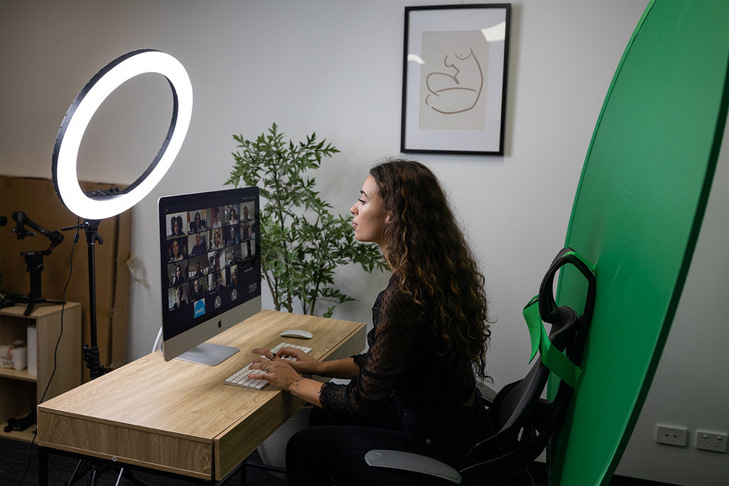 Woman sitting at a desk with her screen visible on Zoom with a green screen attached to chair and a LED ring light directly in front