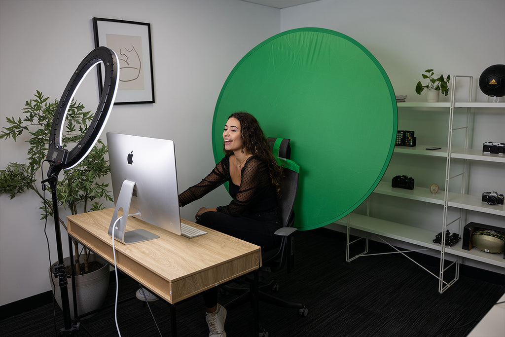 Woman sitting at a desk smiling at a computer with a green backdrop attached to her chair and a large ring light