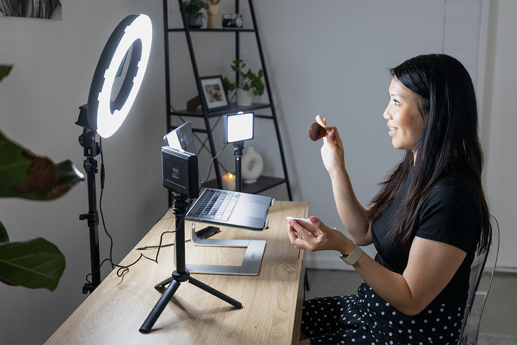 Female sitting at a desk, holding a makeup brush and looking at a ring light