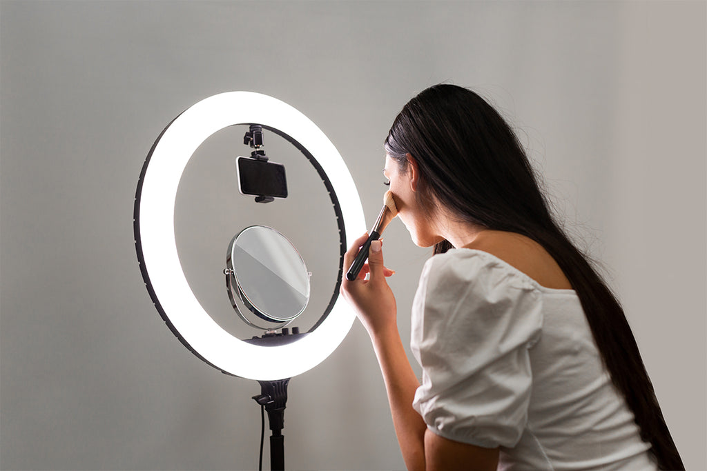 Female with a makeup brush to her cheek and looking at a mirror attached to a ring light