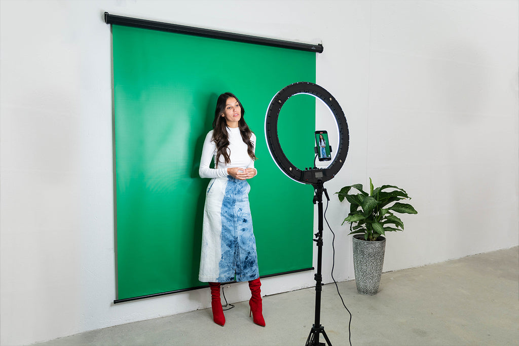 Woman standing in front of a green backdrop looking at a ring light directly in front of her with a phone mounted and recording her