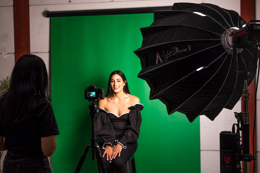 Smiling female model sitting in front of a green paper backdrop on a stand with a camera directly in front of her and two LED side lights