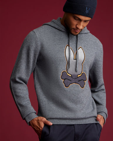 un homme portant un pull en cachemire, pull gris avec un gros logo psycho bunny au milieu. l'homme porte aussi un chapeau gris