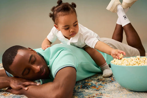 a man laying on the ground with a baby girl on his back trying to take pop corn from a bowl