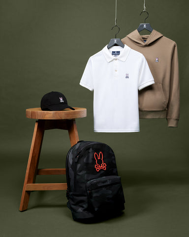 image of a psycho bunny kid polo in white, a backpack, a taupe hoodie hanging and a cap on a chair