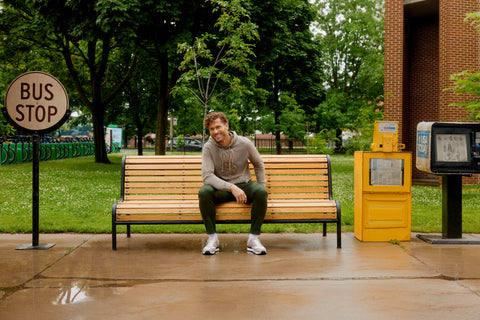 a model with curly hair sitting on a bench with a beige sweatshirt, with a khaki chino and sneakers in a park