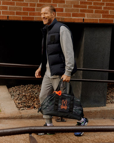 homme marchant avec un sac de week-end dans la main gauche et portant une doudoune sans manches de Psycho Bunny en noir avec un survêtement gris