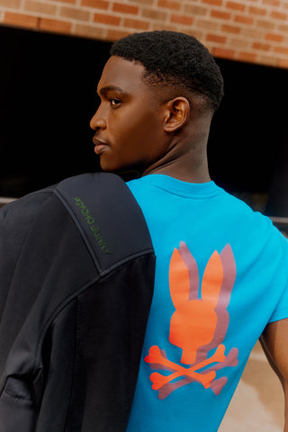 image of a model from the back with a blue psycho bunny back graphic tee in orange and with a coat on his shoulder