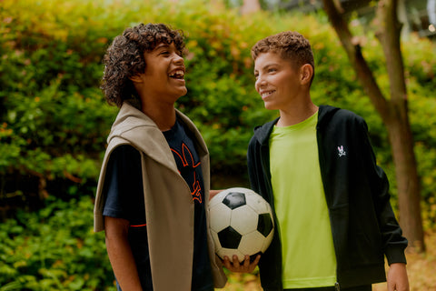 two kids laughing together on with curly hair, a sweater on his shoulder and a soccer ball and the other with a zipped hoodie and a neon yellow tee