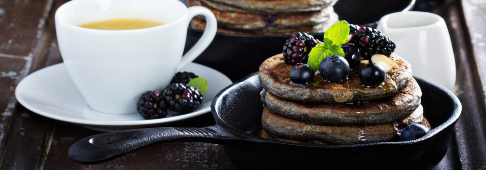 A wooden surface with a cup of coffee and a cast iron skillet with a stack of buckwheat pancakes topped with blackberries, blueberries, and a sprig of mint.