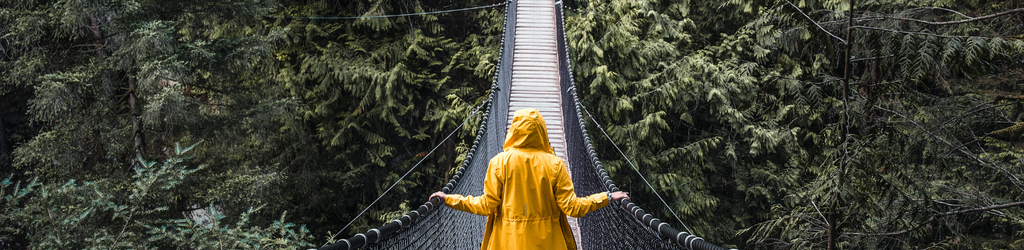A suspended bridge in a wooded area, a person with a yellow raincoat walks across. 