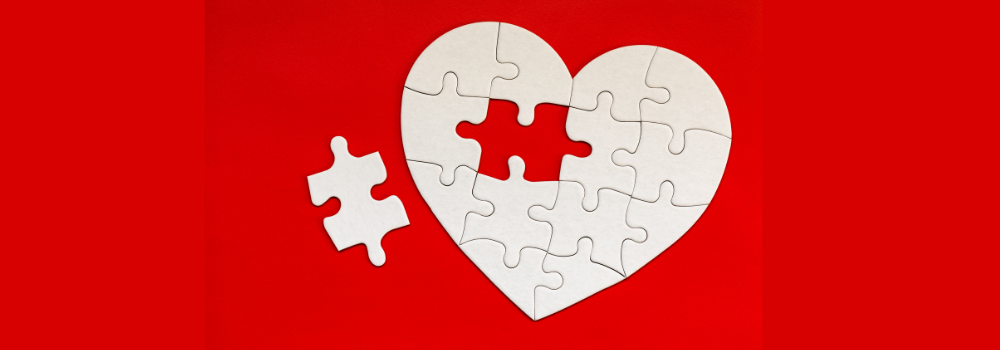 A red background with a white heart made of puzzle pieces; one puzzle piece remains on the red background.