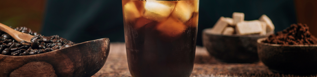 A rustic background depicting the makings of cold brew coffee with the focal point of a glass of cold brew coffee with ice cubes.