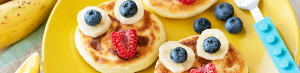 A yellow plate of small pancakes with fruit funny faces made of bananas, blueberries, strawberries, and peanut butter.