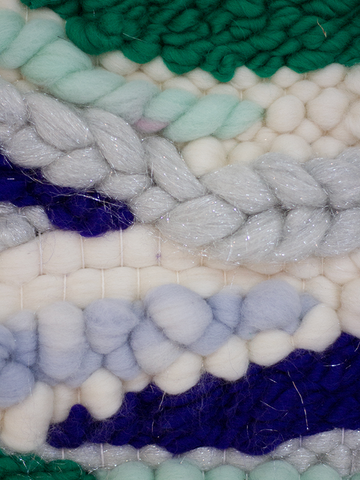 This is a close up of a woven wallhanging in colours of blue, green & natural. The weft yarns used are chunky and include the sumac technique that created the braided shape through the centre