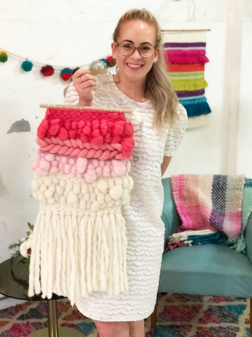 This is a photo of a blonde woman wearing glasses holding a woven wallhanging she has made. The wallhanging features chunky yarns in shades of pink and natural.