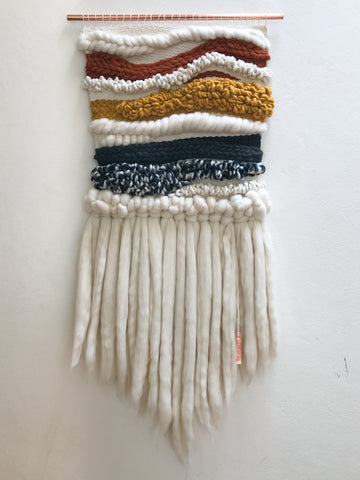 This picture is of a finished woven wallhanging featuring different weaving techniques and various yarns to create texture. The colours are black, natural, terracotta & mustard. this piece was inspired by the carvings and sculptures from the Tiwi Islands