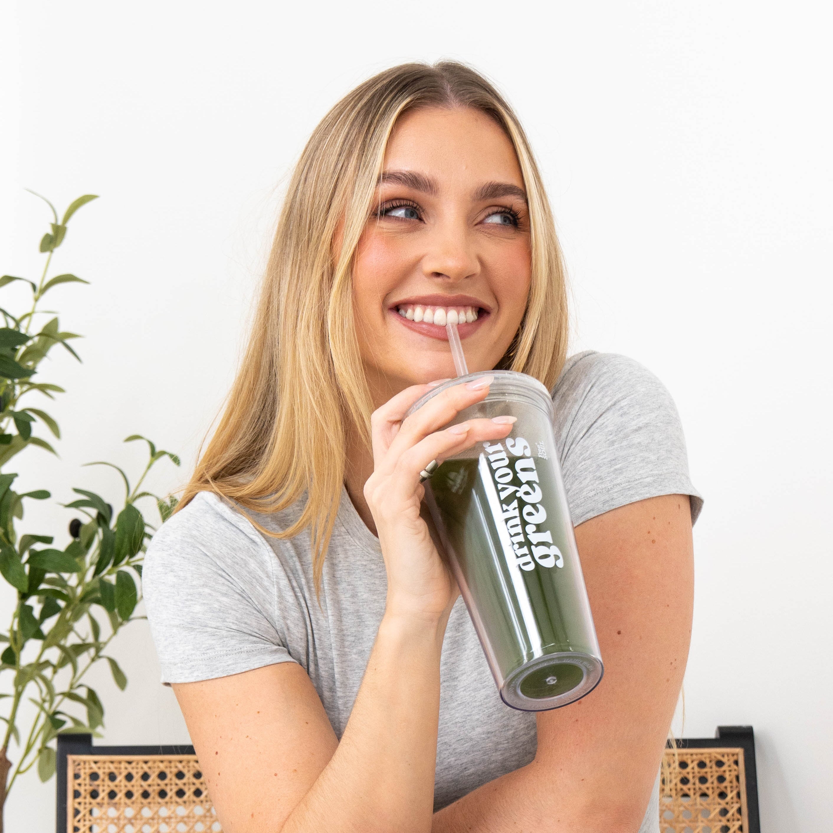 Smiling person drinking a green smoothie from a transparent cup with a straw.
