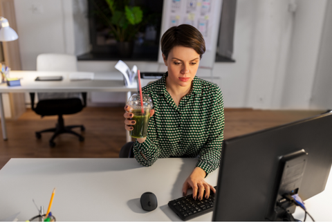 A poised professional woman gracefully sips a green juice at her office desk, focused on her laptop, radiating an aura of concentration and well-being.